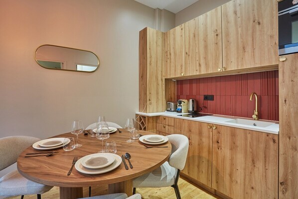 A dining area with a round wooden table set for four, equipped with plates, cutlery, and wine glasses. A kitchen area with wooden cabinetry and a red backsplash is visible in the background.