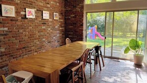 Un comedor con pared de ladrillo, mesa de madera, sillas y un ventanal con vista a un parque infantil.