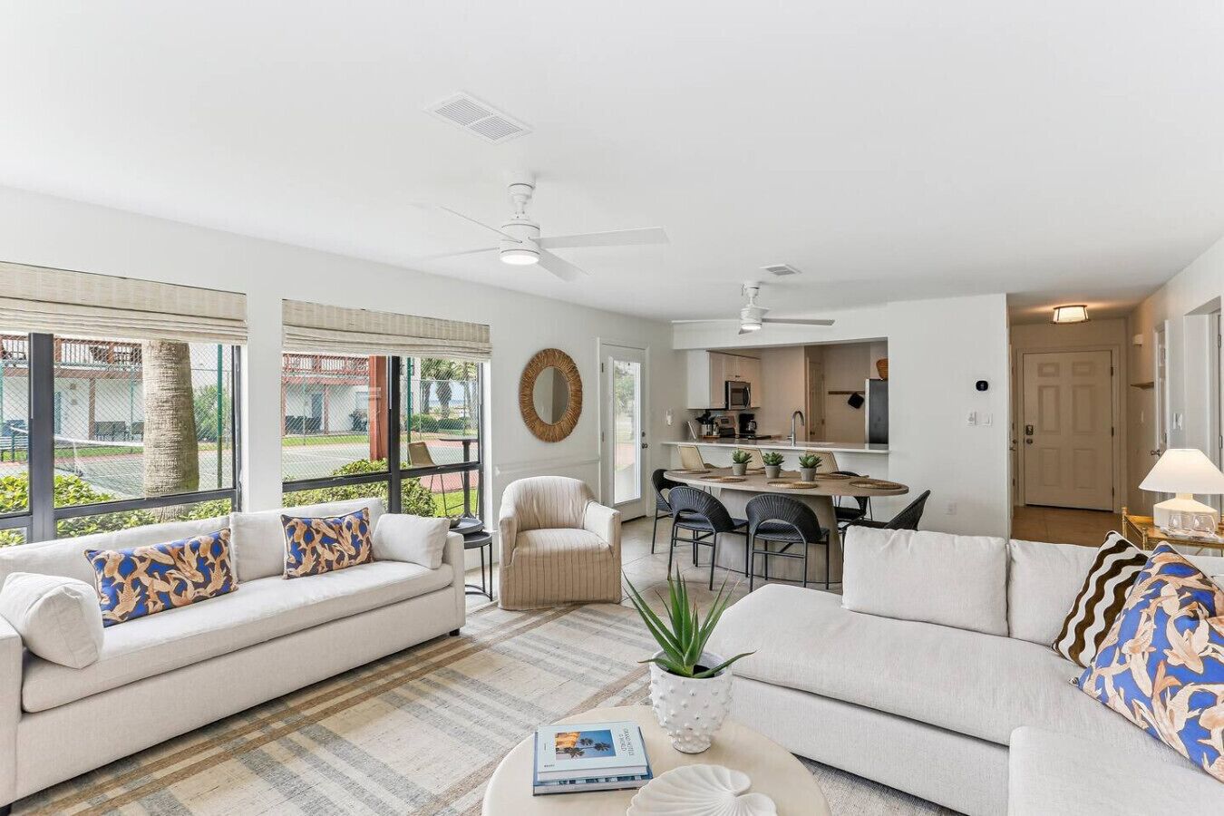 Uma sala de estar ampla com sofá, poltronas e mesa de centro. Há uma área de jantar com mesa e cadeiras, e uma cozinha ao fundo.