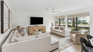 En moderne stue med sofa, lænestole, sofabord og fjernsyn. Der er ventilator, store vinduer og udsigt ud.