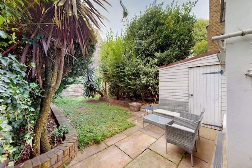Un jardin avec un espace patio, un abri de jardin blanc et un arbre.