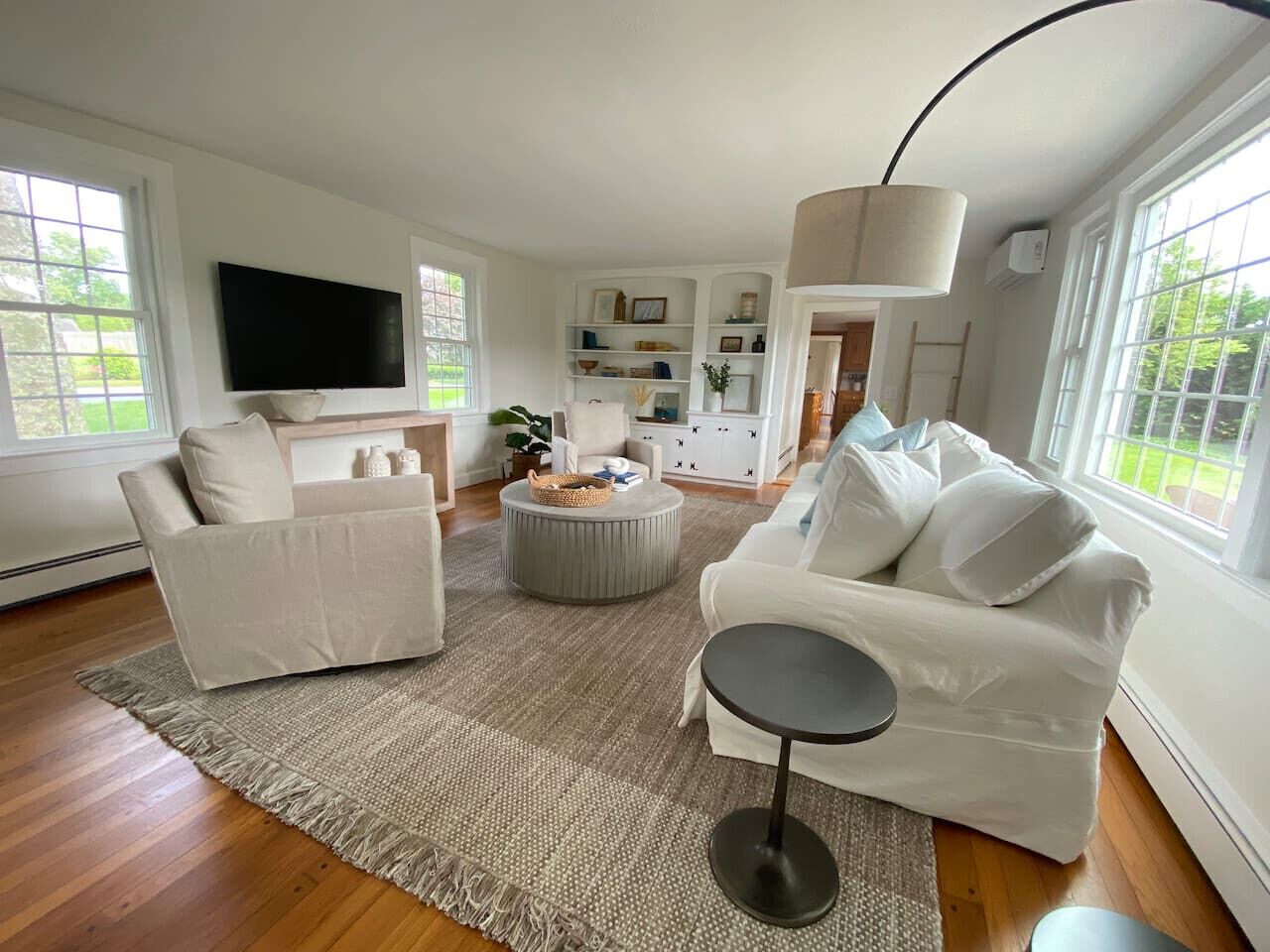Een moderne woonkamer met een grote flatscreen televisie, twee witte banken, een rond poefje en een klein bijzettafeltje.