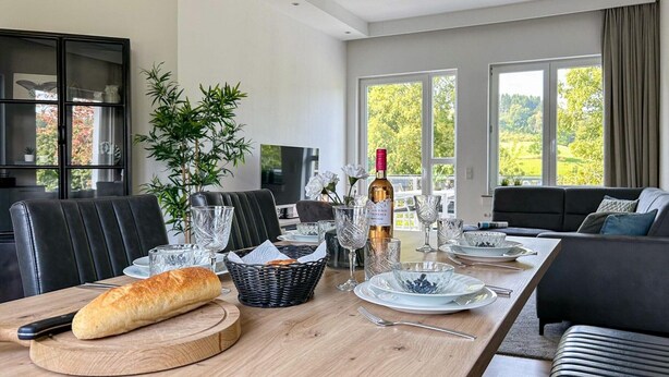 A modern dining area with a wooden table set for a meal, a sofa, and a TV in the background.