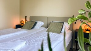 A double bed with white bedding and two pillows, a bedside table with a lamp, and a plant in the foreground.