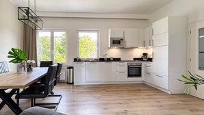 A modern kitchen with white cabinets, a microwave, and an oven. There is a dining table with chairs and a plant on the windowsill.
