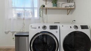 A laundry area with two front-loading washing machines, a trash bin, and a window with sheer curtains. - Spacious 4BEDR/2BATH Getaway w/ King Beds + TVs (Westworth Village)