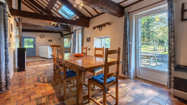 A dining area with a wooden table and chairs, a clock on the wall, and a view of the outdoors through a glass door. - Cottage in a Charming Shepherd’s House, Vast Private Land in the Heart of the Pyrenees (CASTET)