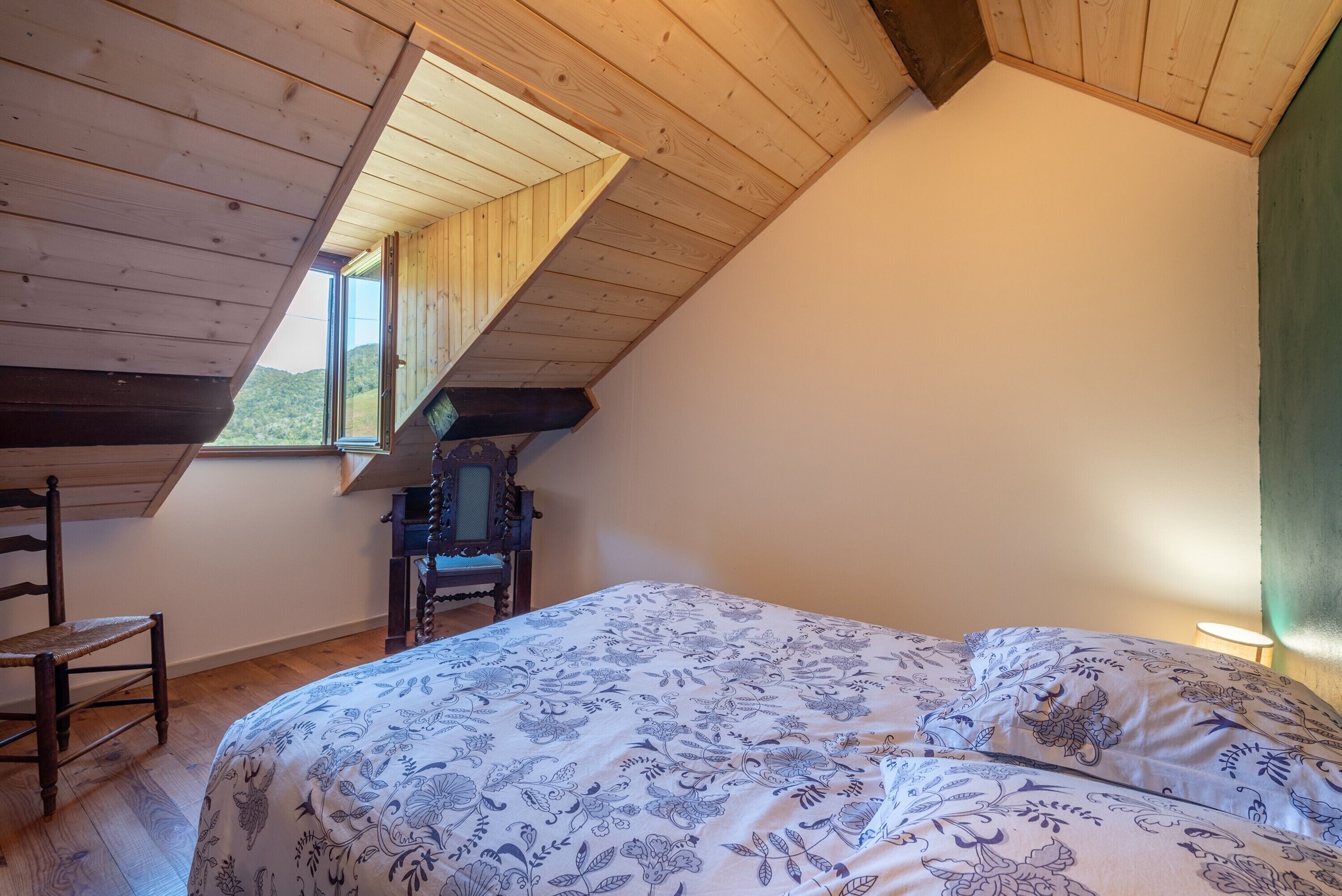 Une chambre avec un plafond en pente, un lit avec une literie à motifs, une chaise et une petite table.