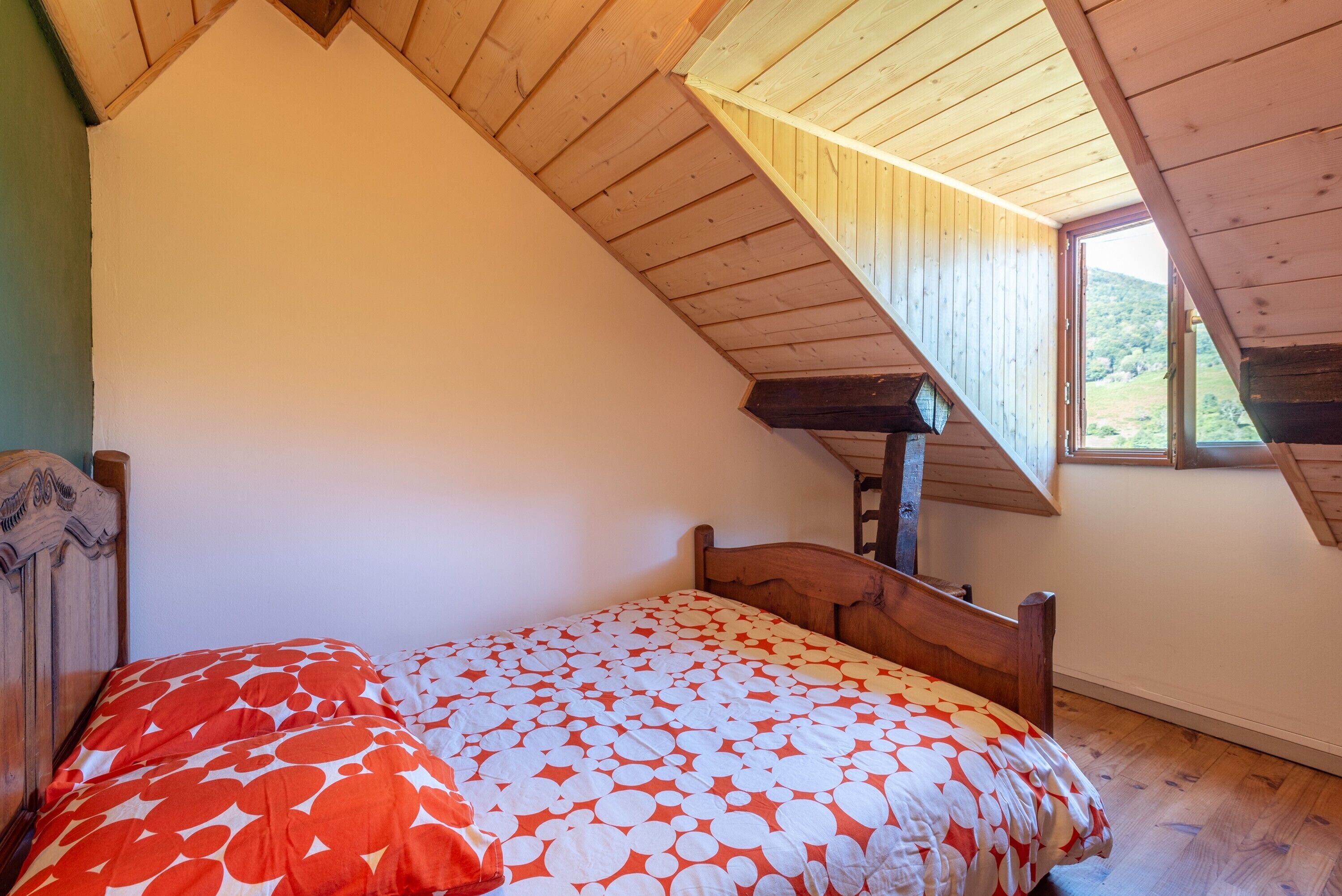 Een slaapkamer met een schuine kap, een houten bedframe en een raam met uitzicht op groen.