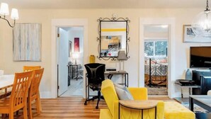 Una sala de estar moderna con un sofá amarillo, una mesa de comedor de madera y sillas. Se ve una silla de oficina negra, una pequeña mesa redonda y un televisor. Un espejo está colgado en la pared, y hay una lámpara sobre la mesa.