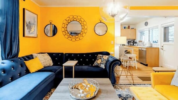 Una sala de estar con un sofá azul, un sillón amarillo y una mesa de centro. Se ve una cocina al fondo.
