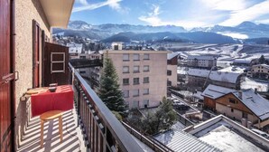 Ein Balkon mit rotem Tisch und Stuhl, mit Blick auf eine Stadt und schneebedeckte Berge im Hintergrund.