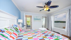 Une chambre avec une couvre-lit rayé aux couleurs vives, un ventilateur de plafond, une vue sur l’eau par la fenêtre et une porte donnant sur l’extérieur.