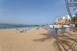 Une plage avec des chaises longues bleues, un abri de plage et un bâtiment doté de balcons.