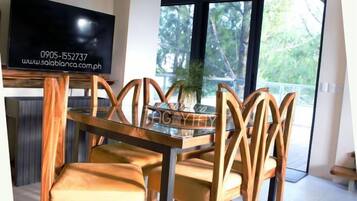 A dining area with a glass table and wooden chairs, a television mounted on the wall, and a view of greenery through large windows.