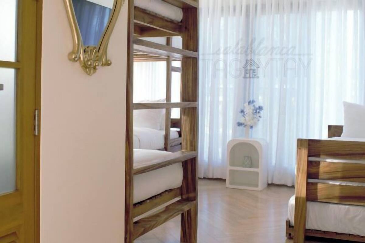 A bedroom with a wooden bunk bed, a white bedside table, and a window with sheer curtains.