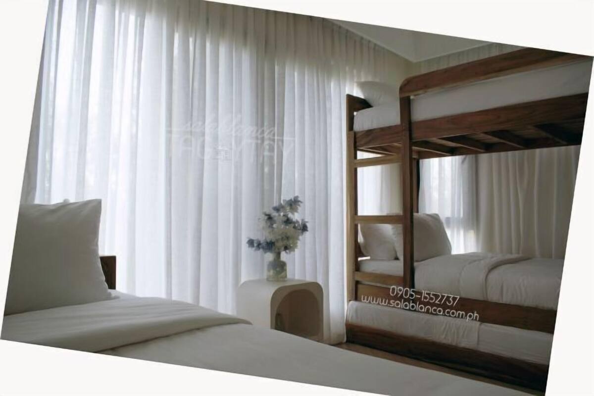 A room with a bunk bed, a bedside table with a vase, and sheer curtains.