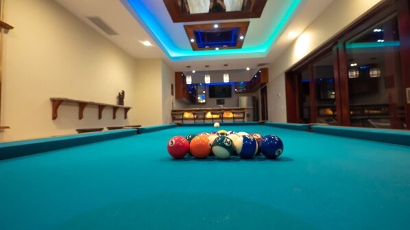 Une table de billard avec les billes alignées, un bar avec des places assises et une vue sur l’extérieur par une fenêtre.