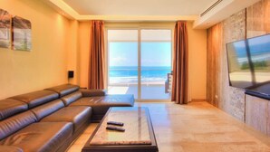 A living room with a sofa, a coffee table, and a TV. There is a large window with a view of the ocean. - Direct Beach Front Ocean View on 7th Floor (Jacó)