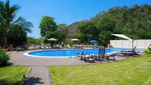 A swimming pool with lounge chairs and umbrellas, surrounded by greenery and a hilly backdrop. - Direct Beach Front Ocean View on 7th Floor (Jacó)