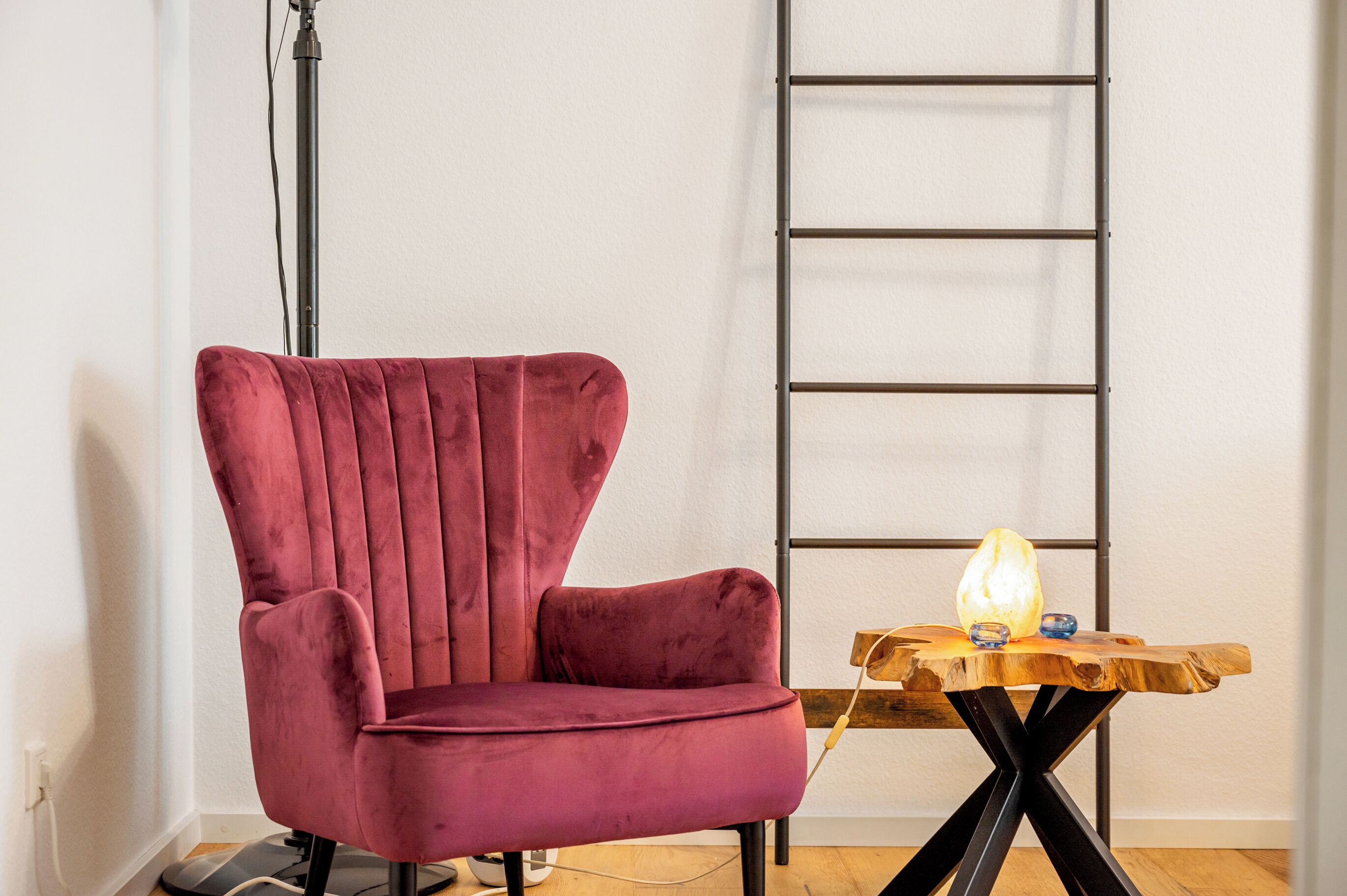 A room with a red velvet chair, a wooden table with a light, and a metal ladder against the wall.