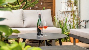 Ein runder Tisch mit einer Weinflasche und zwei Gläsern auf einer Terrasse, im Hintergrund eine Sitzecke und Pflanzen.