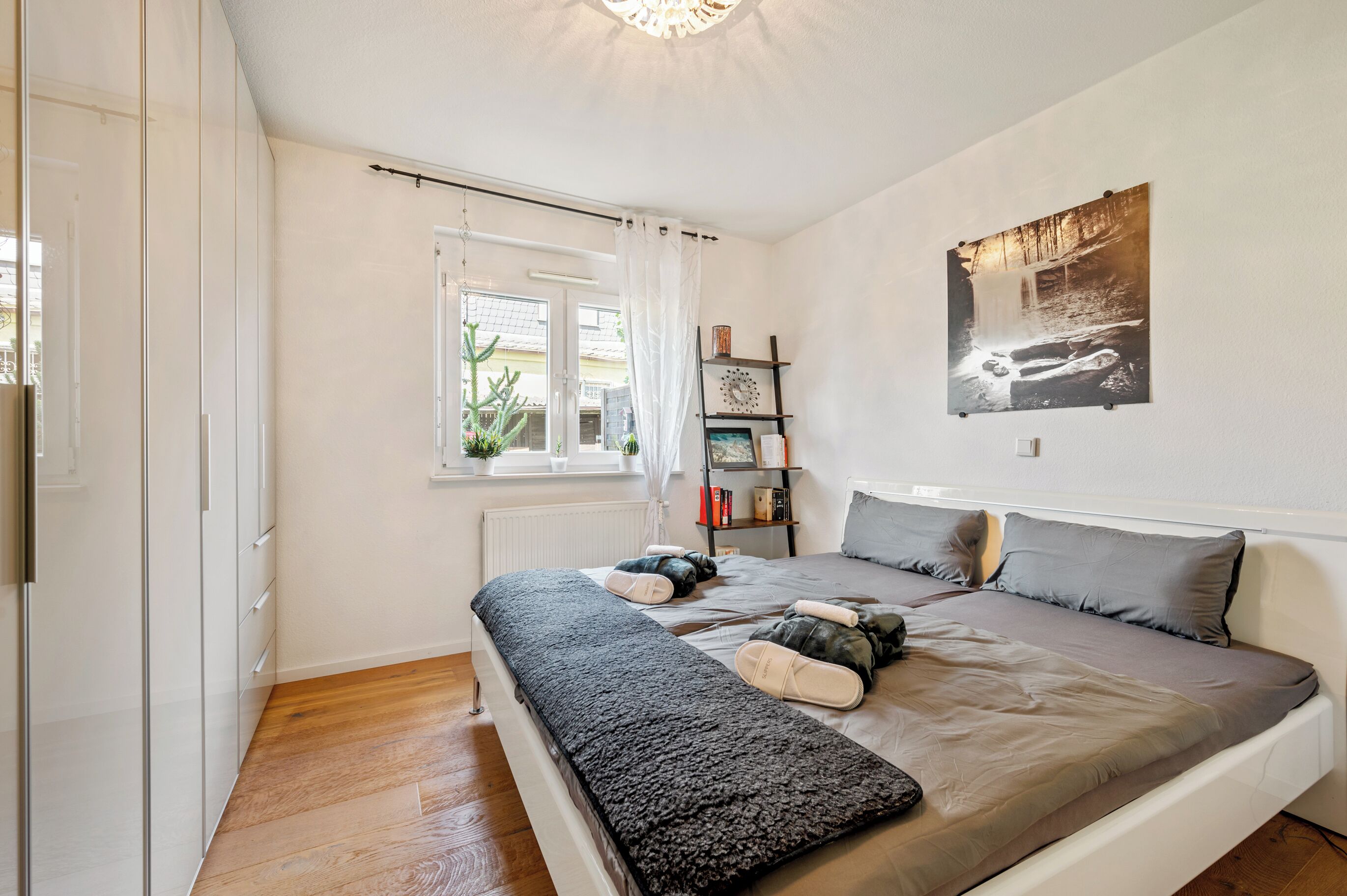 A bedroom with a bed, a window, a radiator, a bookshelf, and a wall-mounted picture.