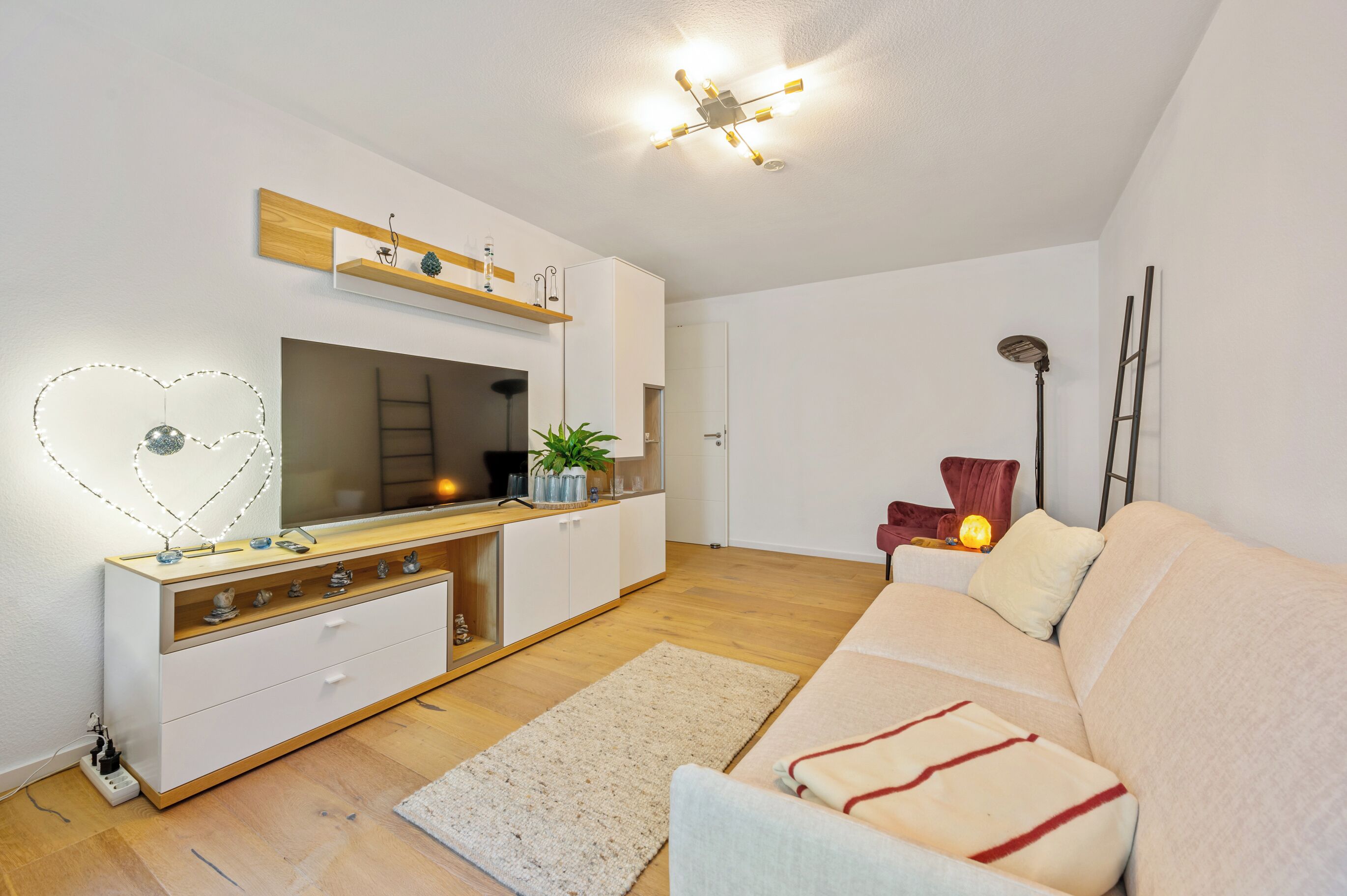 Ein modernes Wohnzimmer mit Sofa, Fernseher, weißem Schrank, Holzregal, Pflanze und Stehlampe.