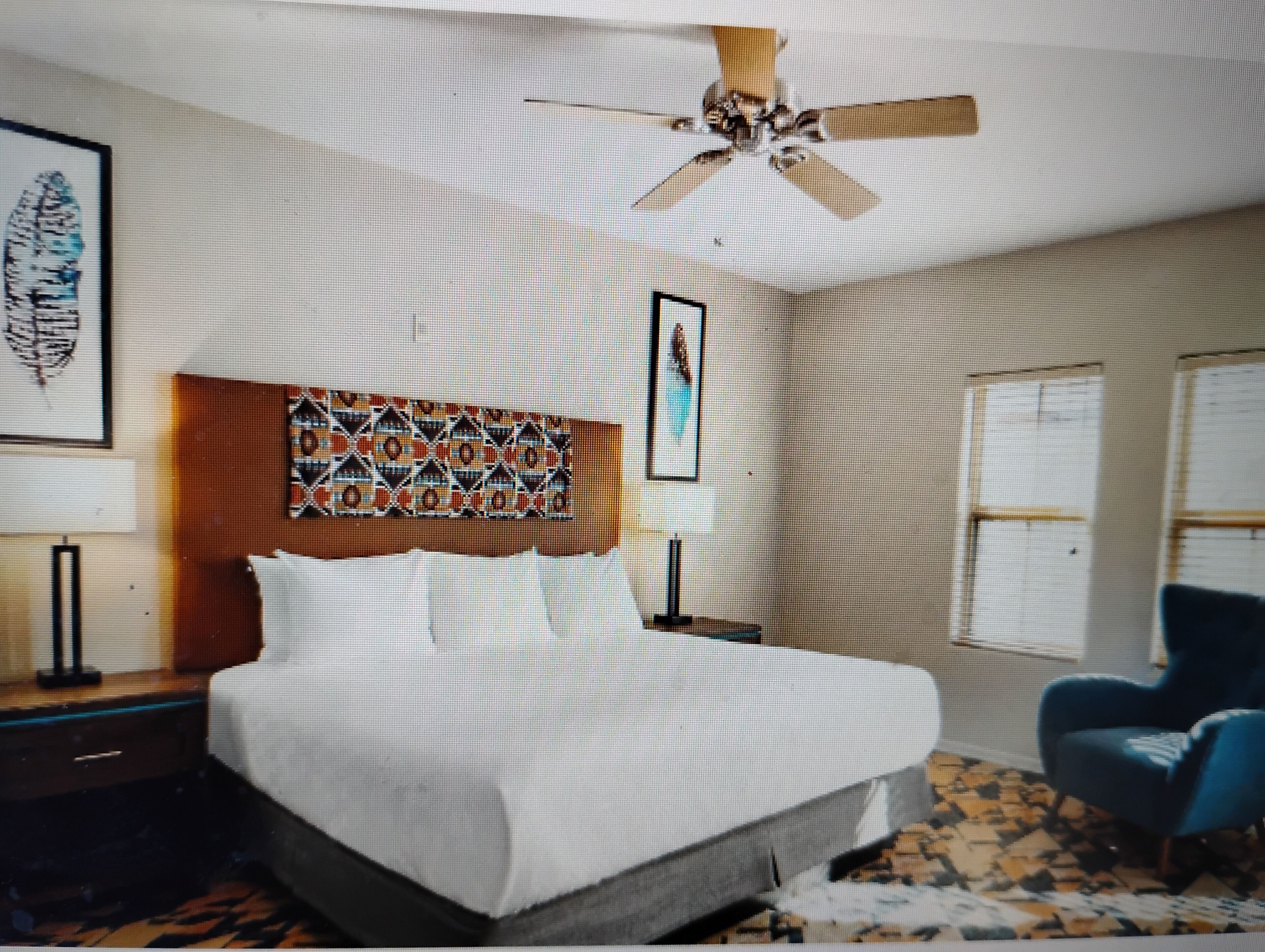 A bedroom with a bed, a ceiling fan, a patterned headboard, a framed picture, a blue chair, and a patterned rug.