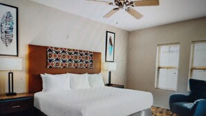 A bedroom with a bed, a ceiling fan, a patterned headboard, a framed picture, a blue chair, and a patterned rug.