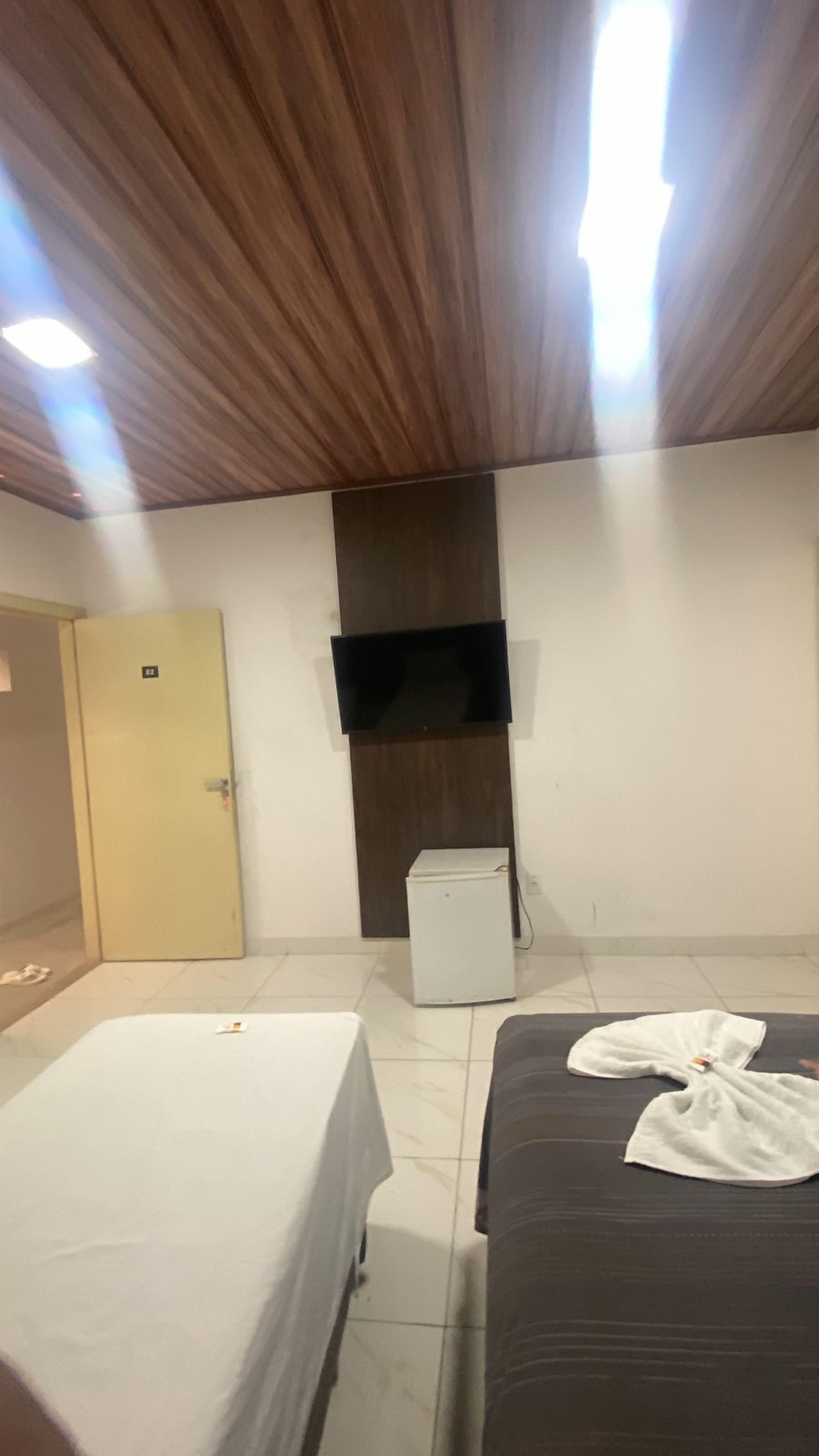 A hotel room with a wooden ceiling, a flat-screen TV mounted on the wall, a small refrigerator, and two beds with white and dark covers.