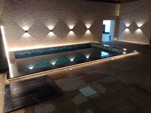 A modern indoor swimming pool with a tiled floor and walls, surrounded by a brick wall with recessed lighting.