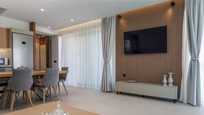 Una habitación de hotel moderna con comedor, televisor montado en una pared de paneles de madera y un ventanal con cortinas.