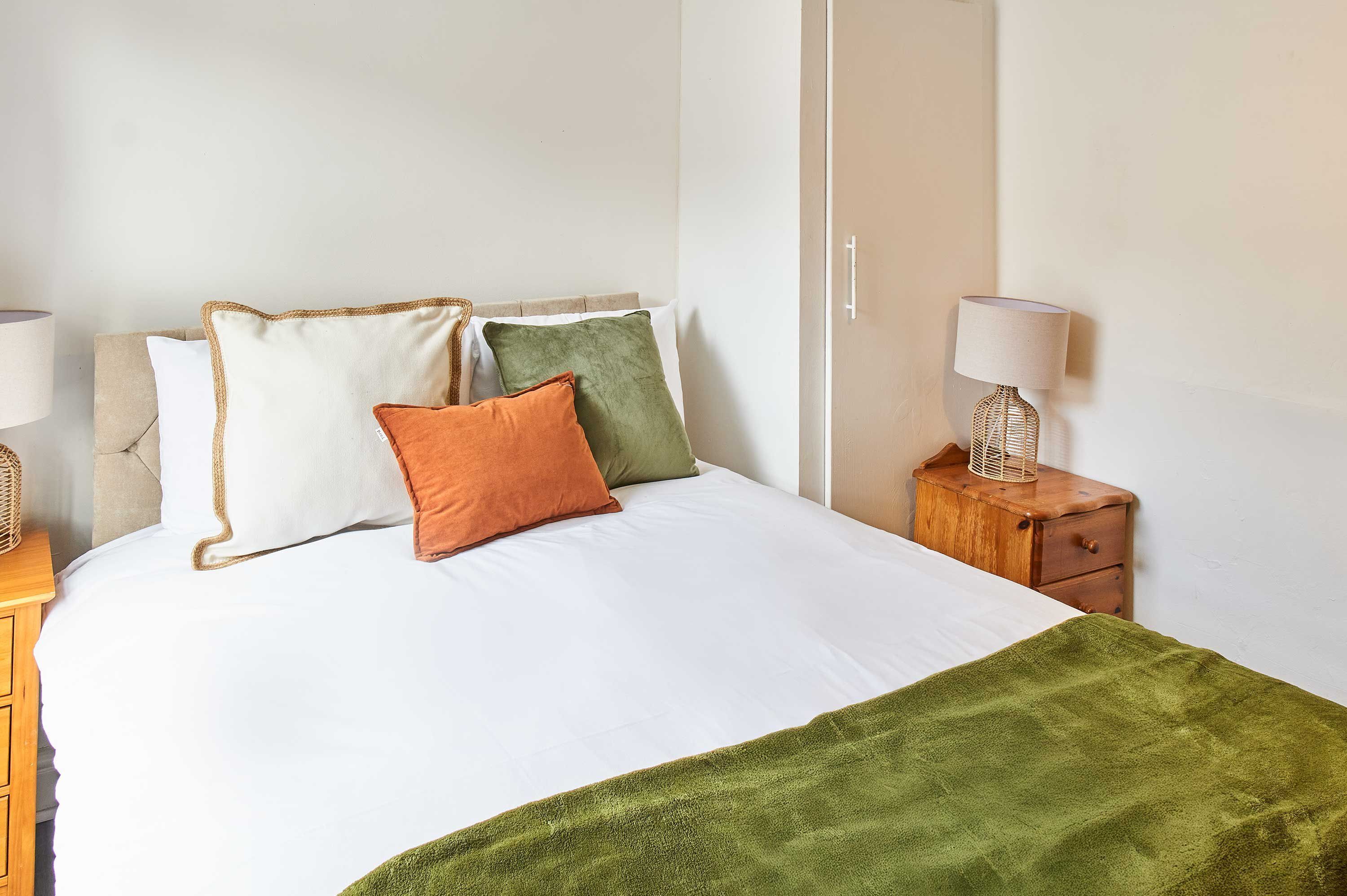 A neatly made bed with white bedding and a green throw blanket, flanked by pillows in various colors, and a wooden nightstand with a lamp beside it.