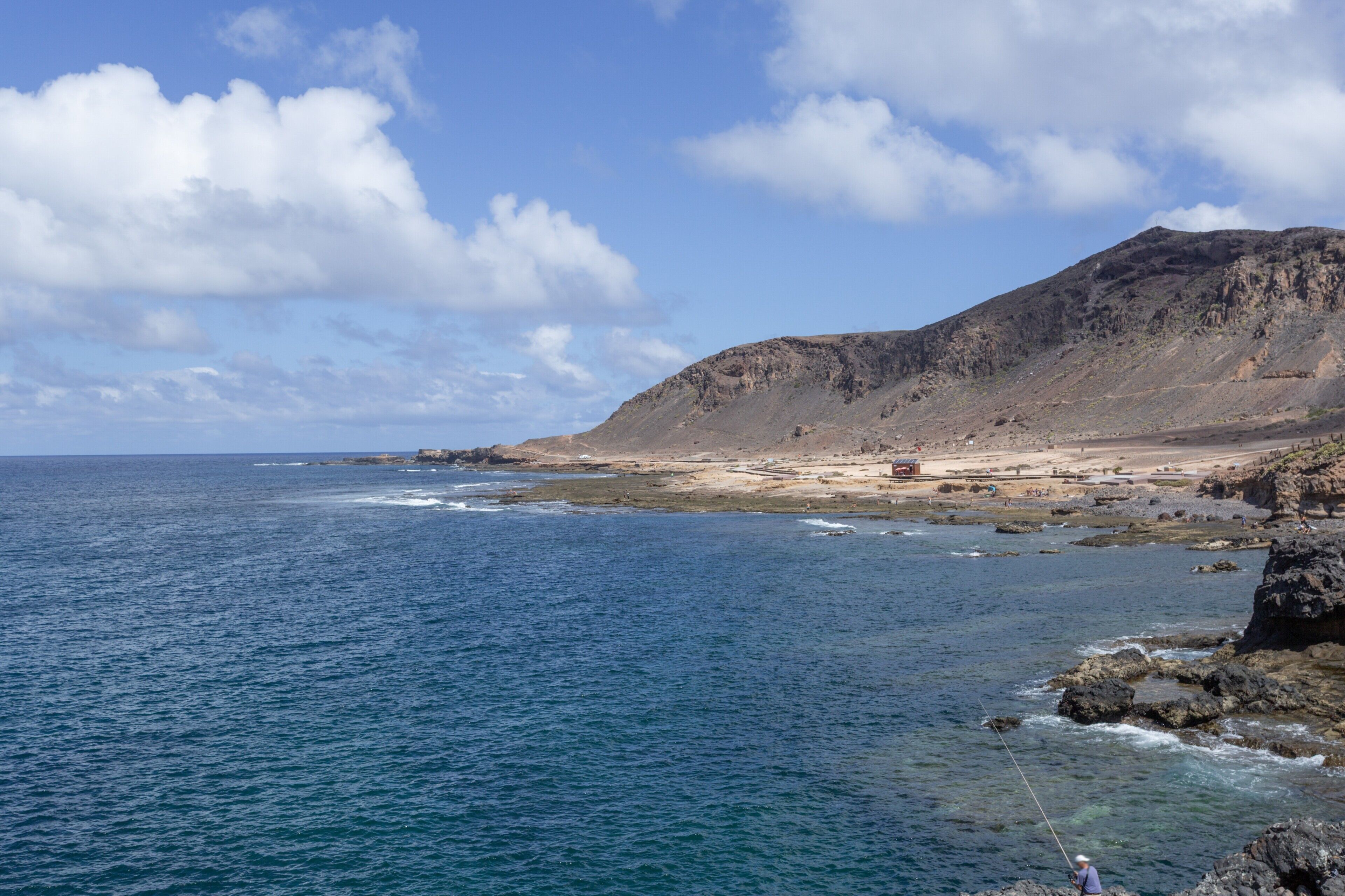 A coastal landscape with a rocky shoreline, a person fishing, and a distant mountain.