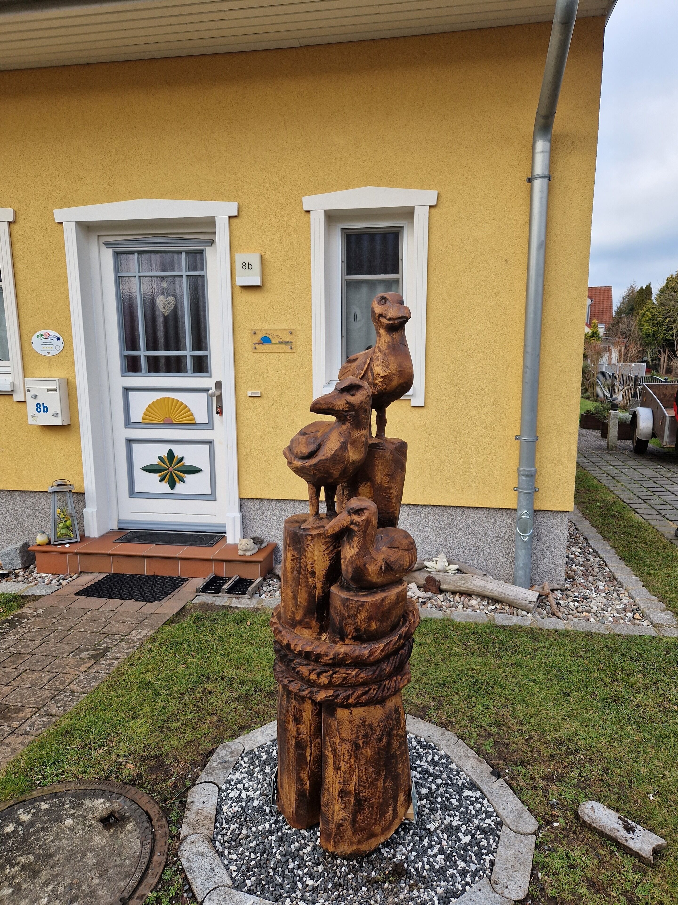 A wooden sculpture of a bird on a pedestal in front of a yellow building with a white door and windows.