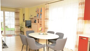Un comedor con una mesa redonda y seis sillas, un televisor, una estantería y una zona de cocina con armarios rojos.