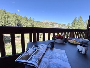A table with a magazine, a pair of glasses, a bowl of fruit, a mug, and a plate of croissants on a balcony overlooking a forested hillside.