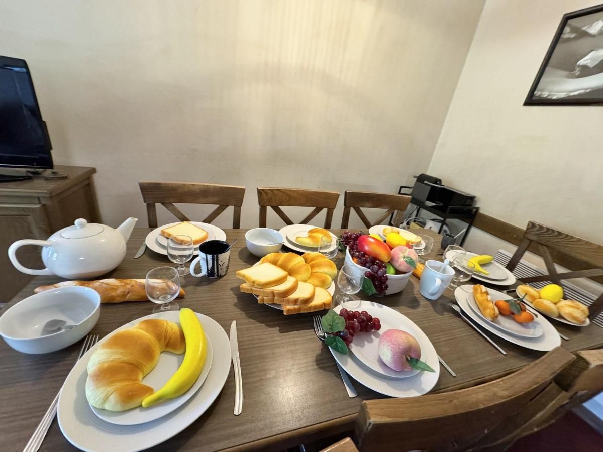 Una mesa de comedor dispuesta con diversos productos para el desayuno, incluyendo pan, frutas y una tetera.