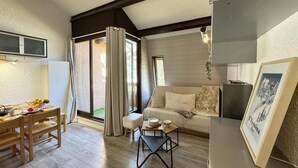A cozy living room with a sofa, a dining table with chairs, and a small coffee table. There is a window with curtains, a framed picture on the wall, and a ceiling with exposed beams.