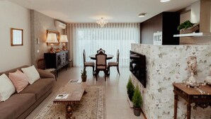A living room with a sofa, a dining table, and chairs. There is a kitchen area with a microwave and a shelf with plants.