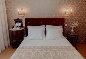 A neatly made bed with a red headboard, two wall sconces, a small round table with a mirror, and a vase with flowers.