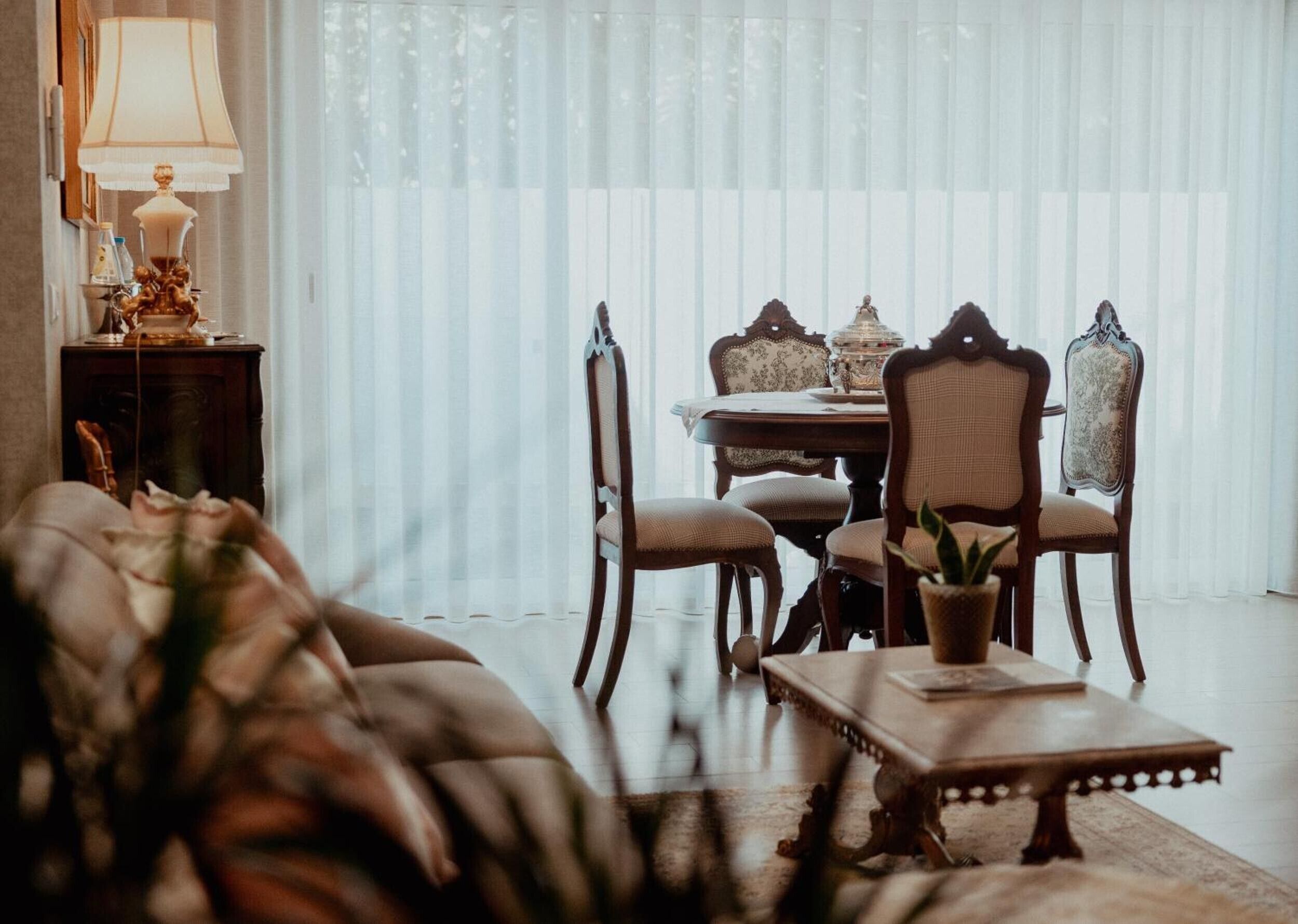 A dining area with a table set for four, surrounded by ornate chairs, a sofa, and a lamp on a side table.