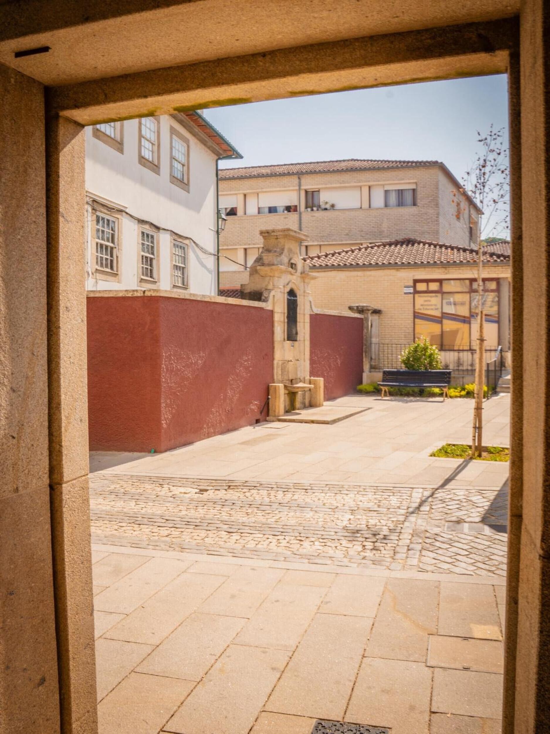 Une porte ouverte donne sur une cour intérieure, où l’on trouve un chemin pavé de pierres, un banc et un petit arbre.