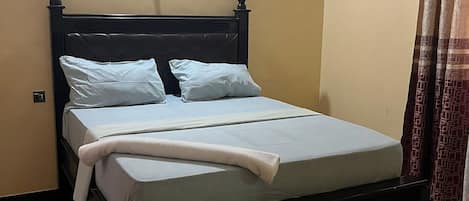 A neatly made bed in a room with a plain wall and a ceiling light.