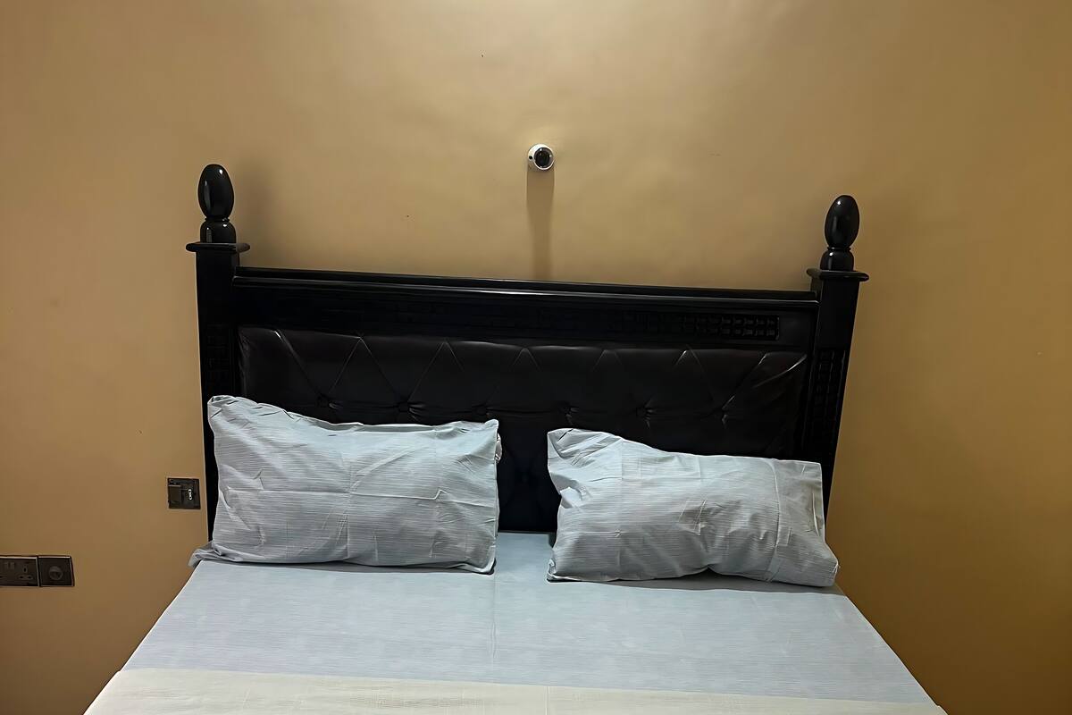 Uma cama bem arrumada com lençóis e almofadas brancos, uma cabeceira preta e um interruptor de luz na parede.
