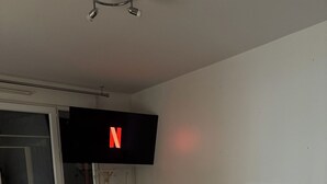 A modern living room with a large flat-screen TV mounted on the wall, a beige sofa with black cushions, a white coffee table, and a small wooden side table. - Private room - Garden & terrace (Noisy-le-Sec)