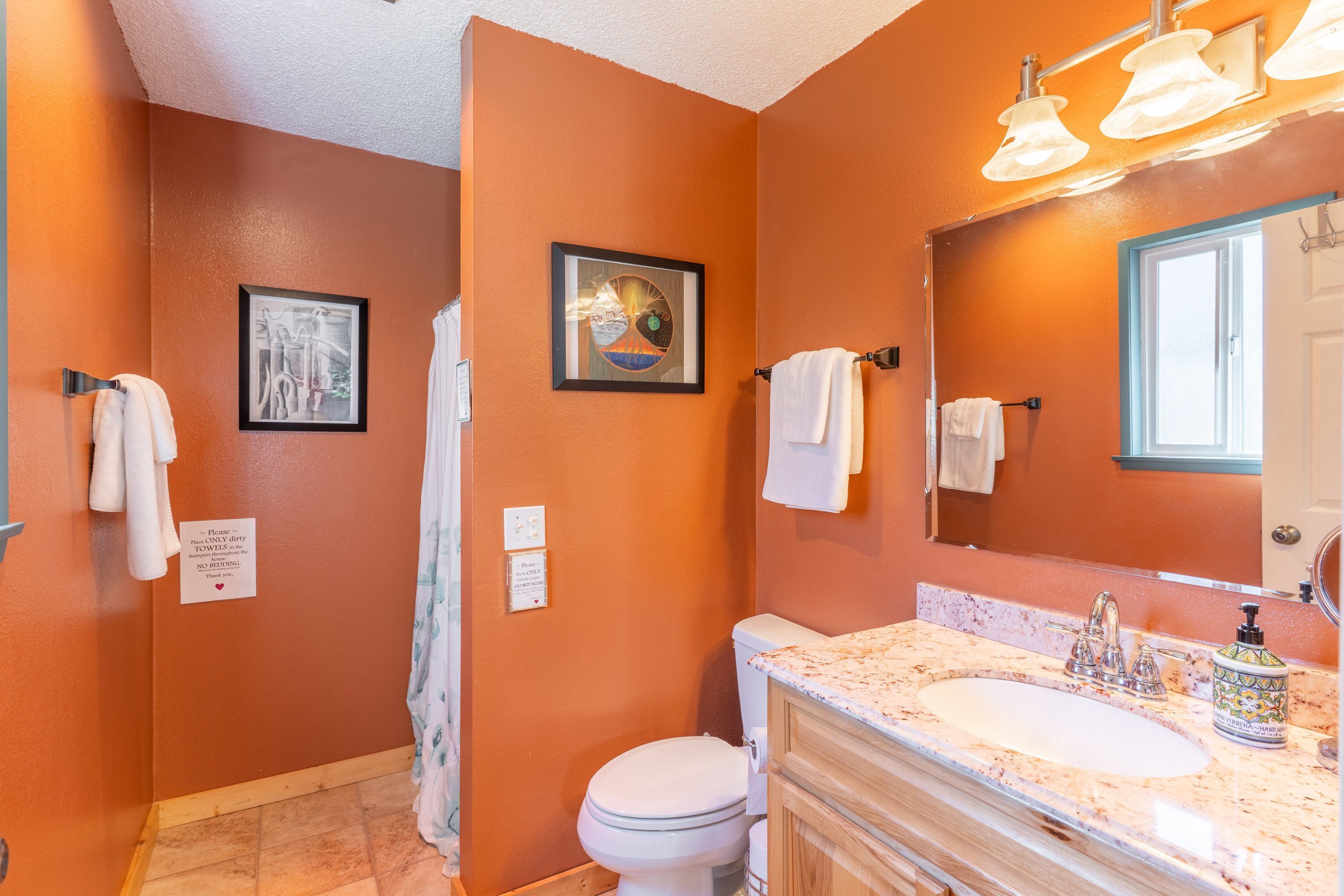 Une salle de bain avec un mur orange, des toilettes blanches, un lavabo avec un plan de travail en marbre et un miroir. Deux tableaux encadrés sont accrochés au mur, et il y a un porte-serviettes avec des serviettes.