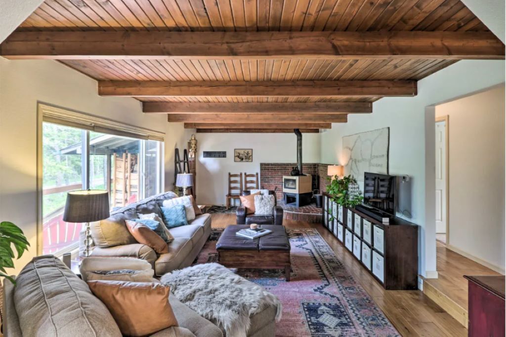 Ein gemütliches Wohnzimmer mit Holzbalkendecke, Kamin, Fernseher und großem Fenster.