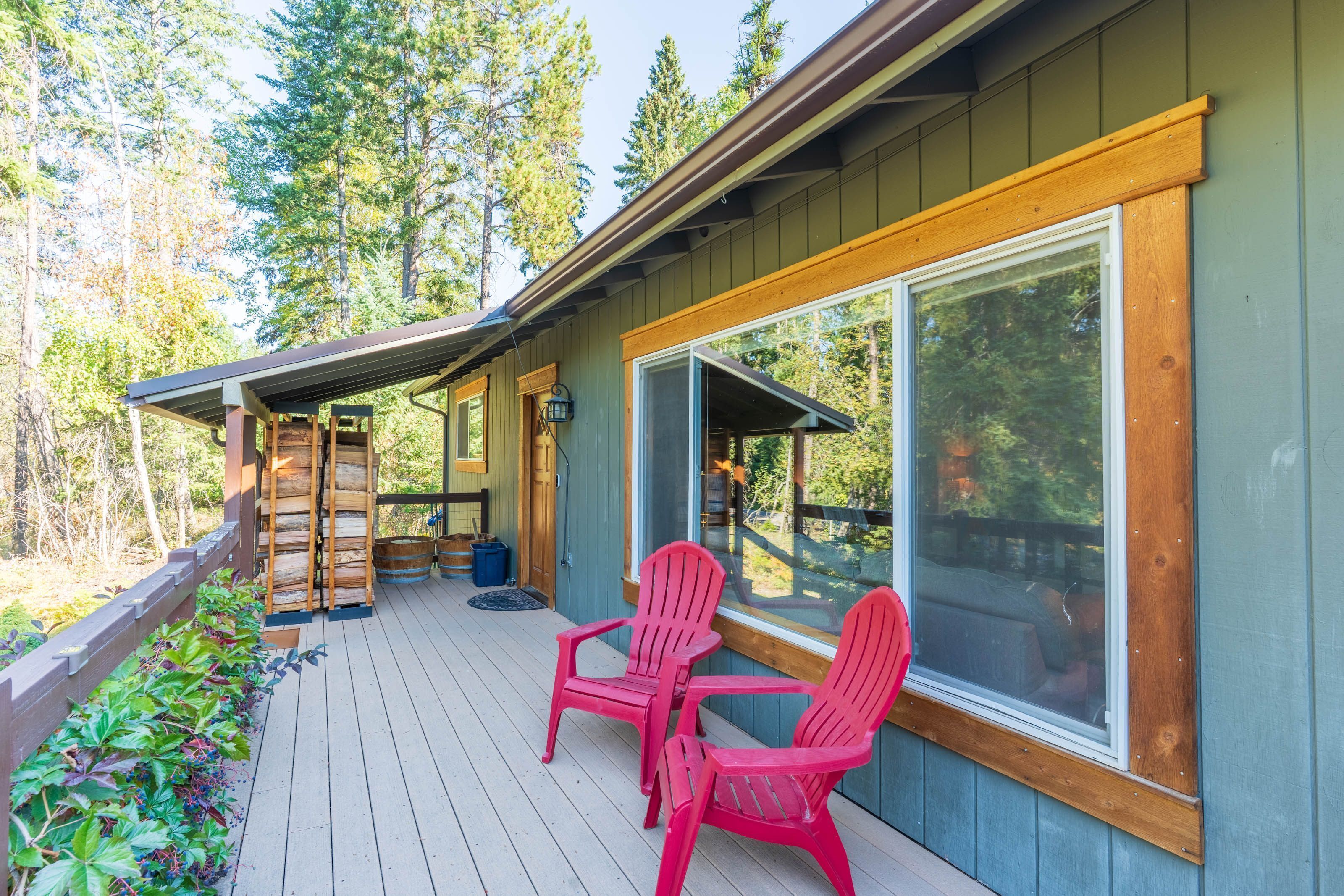 A wooden deck with two Adirondack chairs, a covered area with a wooden bench, and a large window overlooking a forested area.